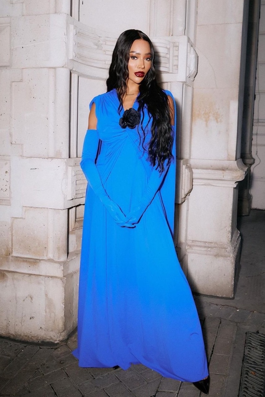 Elegant woman in a flowing bright blue gown and matching opera gloves poses against a detailed stone wall in soft, natural light.