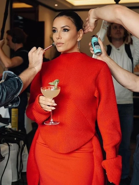 Eva Longoria at a photoshoot with makeup artists around her. She is holding a cocktail and is wearing a red sweater and dress.