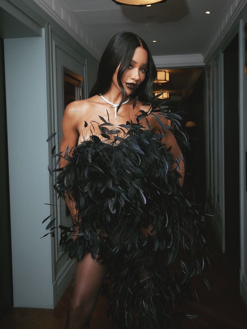 Glamorous woman in an elegant, strapless black feather dress and long dark hair poses in a hallway with gray walls and warm lighting.