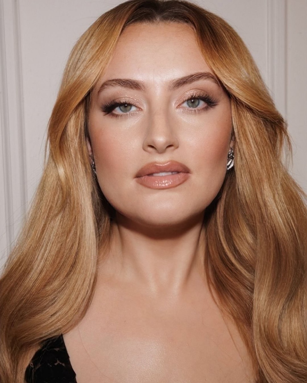 Close-up portrait of an adult female with long, wavy blonde hair, wearing makeup and earrings, in a neutral indoor setting with soft lighting.