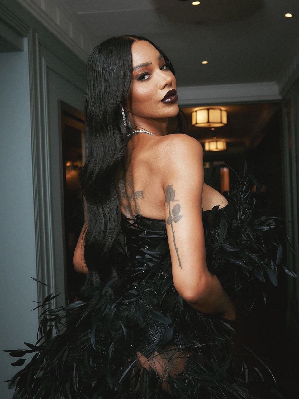A woman in an elaborate black feathered dress looks back over her shoulder, her hair styled straight and dark, set against a neutral interior.