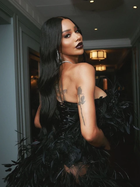 A woman in an elaborate black feathered dress looks back over her shoulder, her hair styled straight and dark, set against a neutral interior.