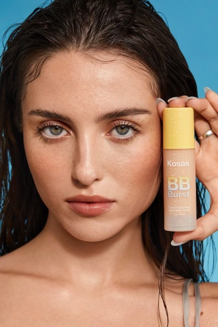 Close-up shot of a fair-skinned woman with brown hair holding a bottle of Kosas BB Burst, against a light blue background.