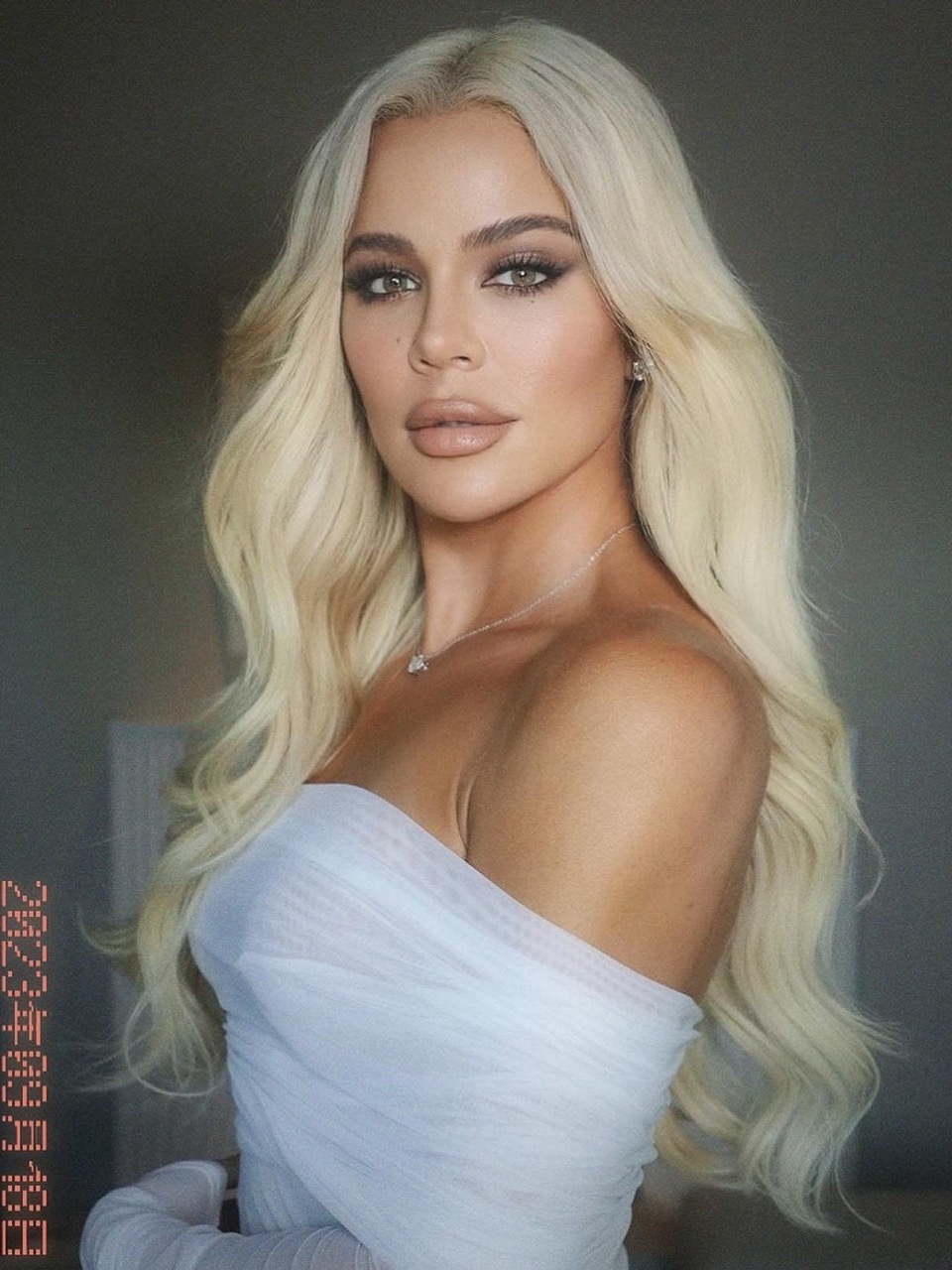 Portrait of a woman with platinum blonde wavy hair, wearing a white strapless dress and jewelry, posing indoors against a grey background.