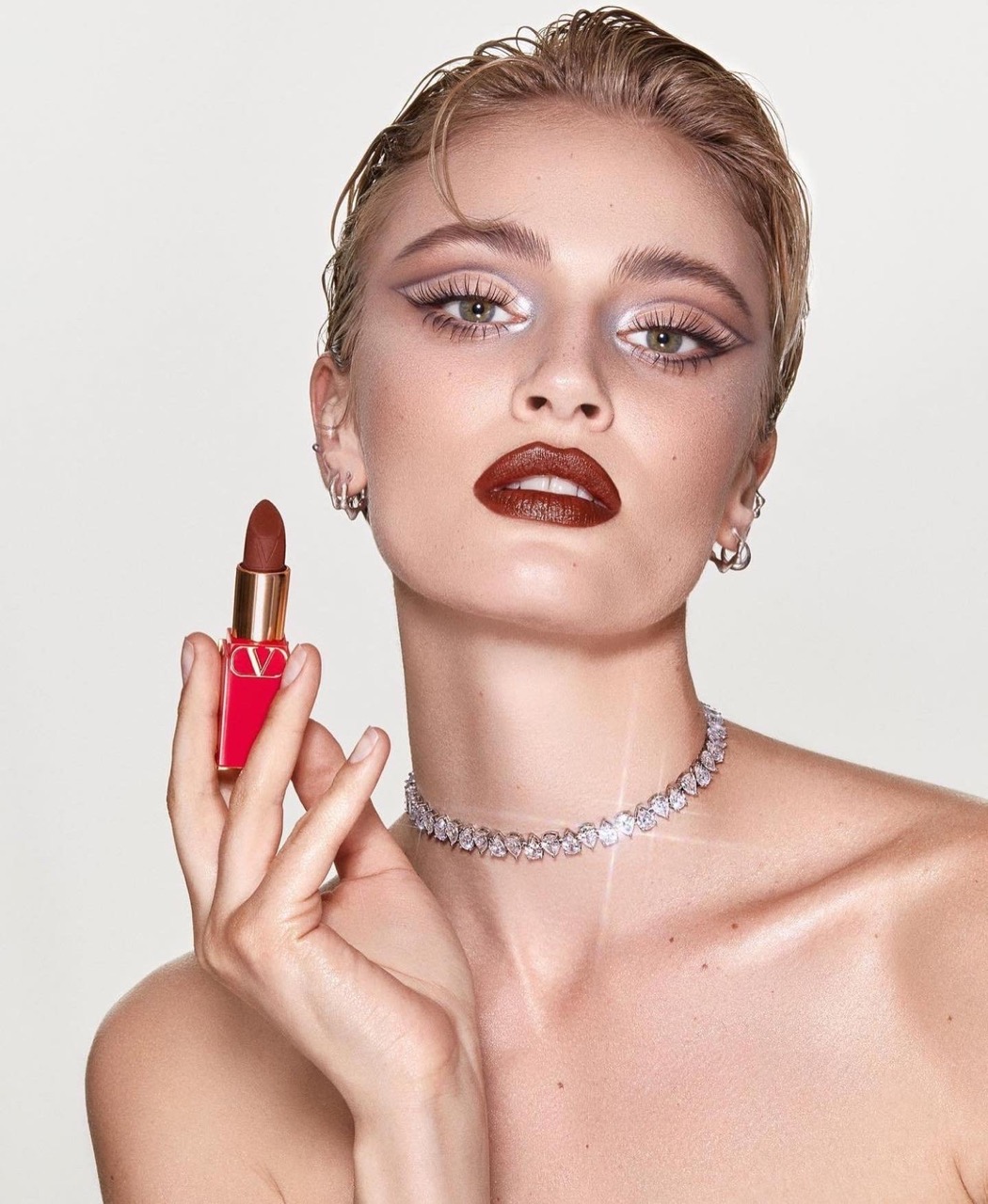 Blonde-haired model in a beauty shot holding lipstick tube, wearing bold eye makeup, silver jewelry, and dark lipstick against white background.