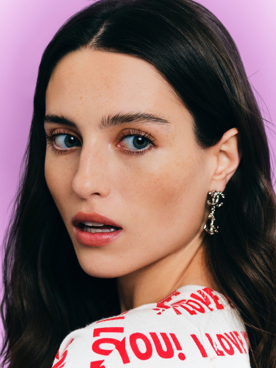 Close-up portrait of a woman with dark hair, blue eyes, and silver earrings against a purple background.