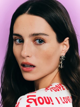 Close-up portrait of a woman with dark hair, blue eyes, and silver earrings against a purple background.