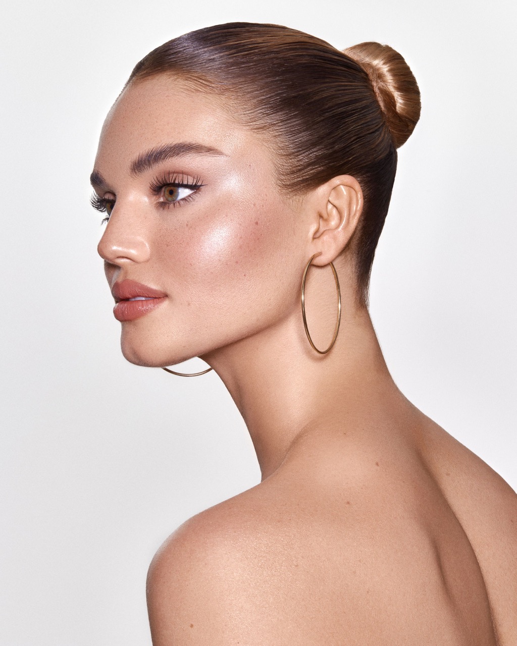 Close-up portrait of an adult woman with tanned skin, featuring gold hoop earrings and a hair bun, against a plain white backdrop.