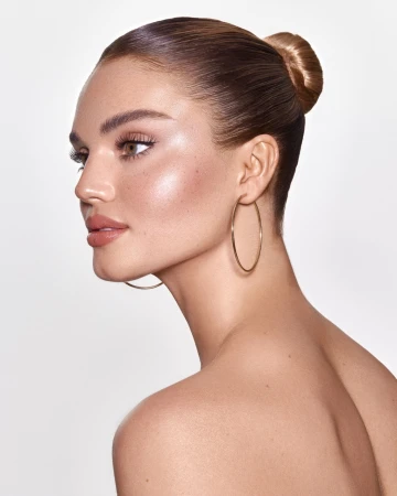 Close-up portrait of an adult woman with tanned skin, featuring gold hoop earrings and a hair bun, against a plain white backdrop.