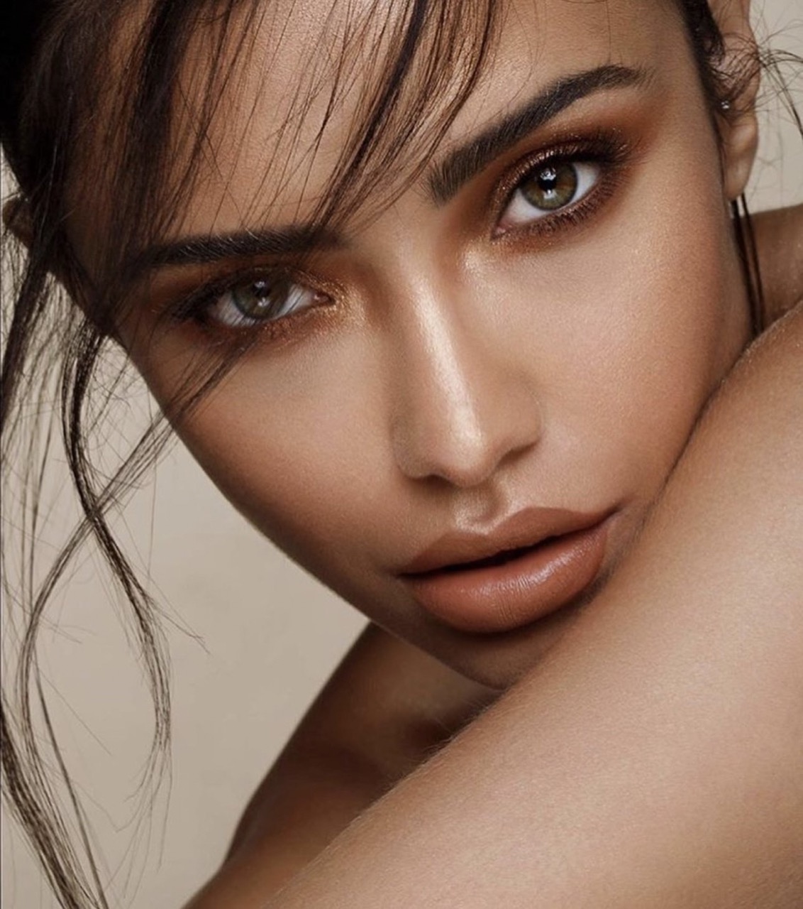 Close-up portrait of a woman with striking makeup and loose dark hair, posing in a beauty or fashion context, set against a neutral background.