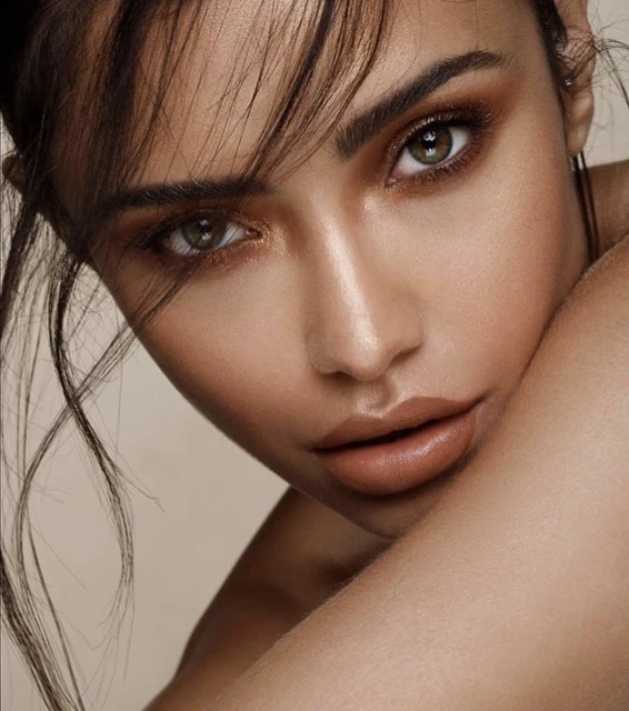 Close-up portrait of a woman with striking makeup and loose dark hair, posing in a beauty or fashion context, set against a neutral background.