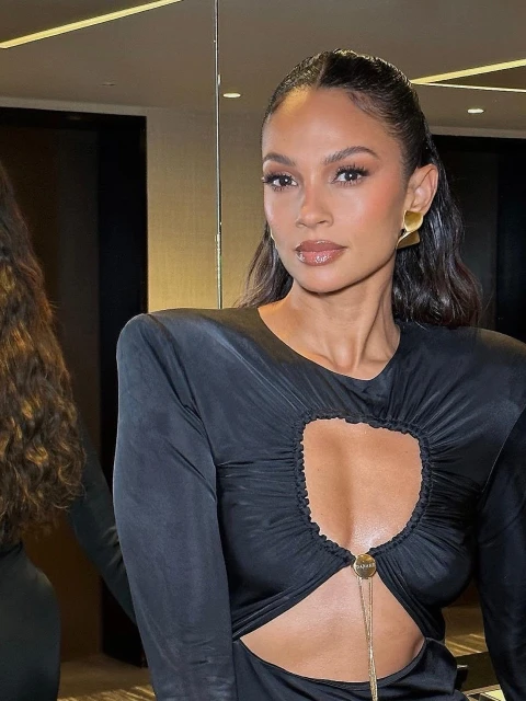 Close-up portrait of a woman with dark hair wearing a black long-sleeve dress with a chest cut-out and large gold earrings indoors.