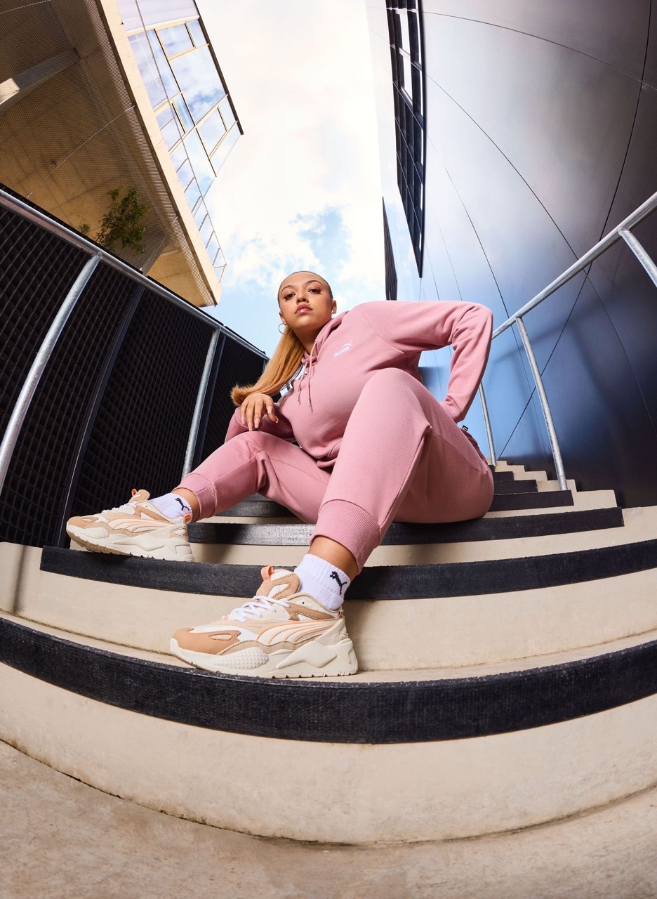 Woman in a pink Puma tracksuit sitting on striped outdoor stairs. She wears cream and tan sneakers with a building background.