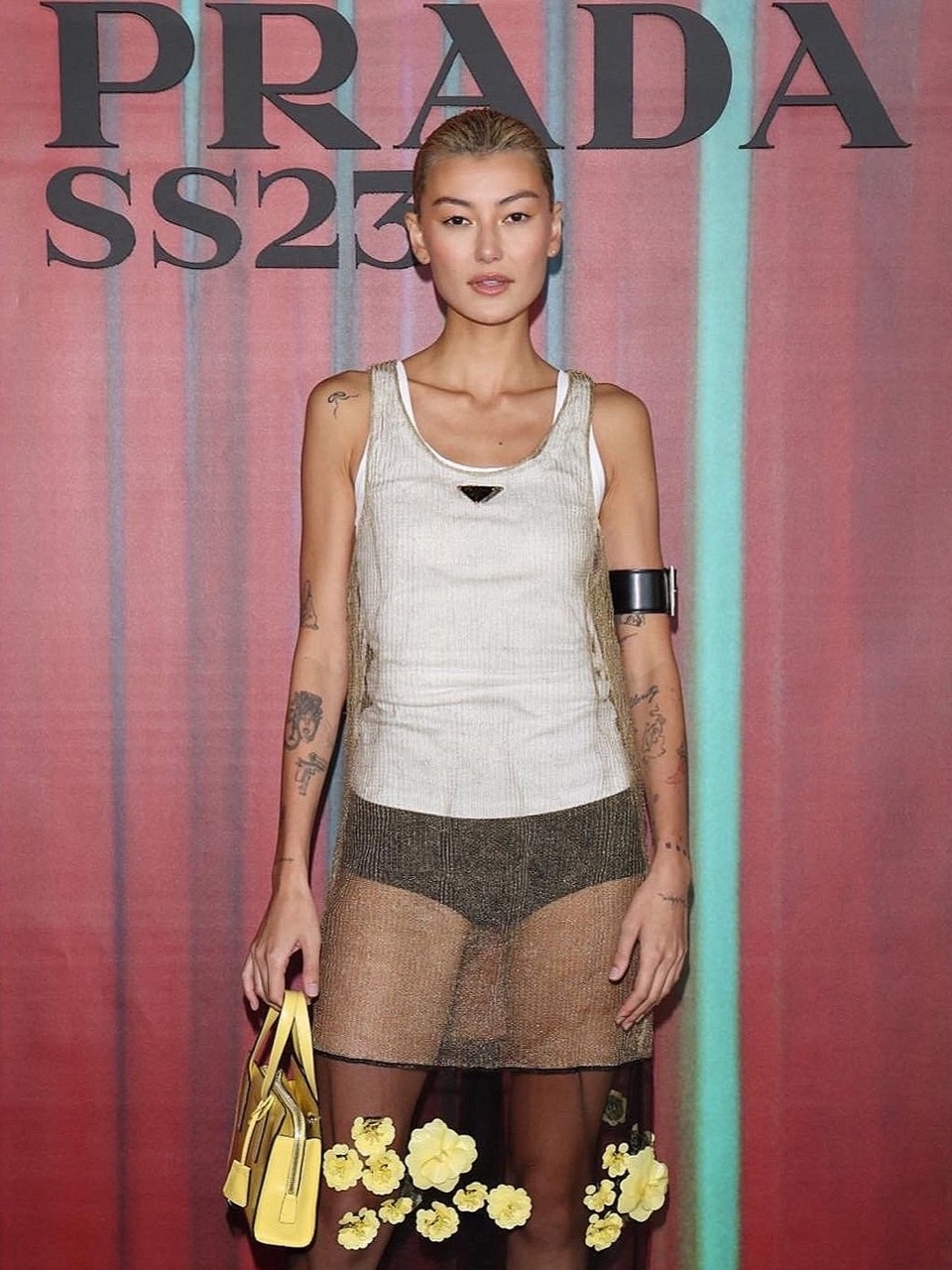 Woman with fair skin and blonde hair poses with a yellow handbag in front of a backdrop with the Prada logo and SS23 text.