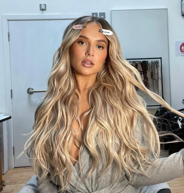 Blonde woman with hair clips, styled wavy hair, and grey wrap top poses in studio environment looking at camera. Beauty portrait.