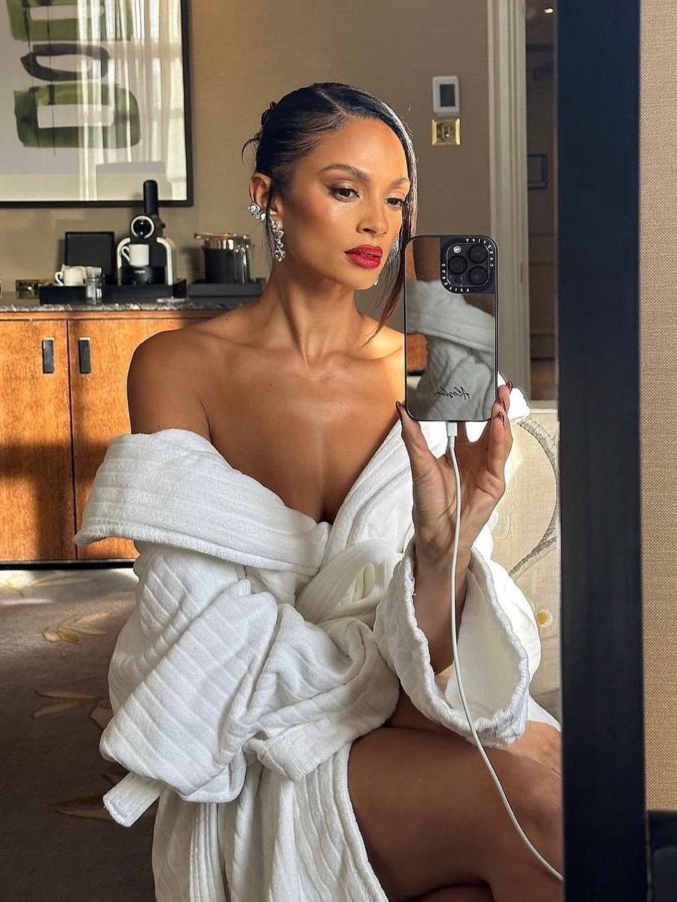 Adult female with bare shoulders and makeup, posing in a white robe while holding a phone in a neutral-toned hotel room setting.