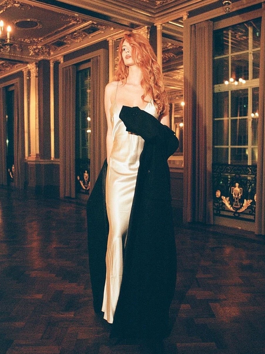 Elegant woman with red hair in white dress and black coat stands in ornate interior with chandeliers and mirrored walls, posing gracefully.