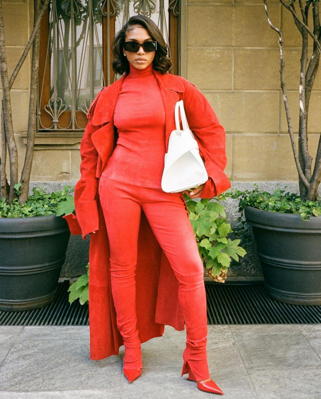 Woman in vibrant red monochrome outfit holding a white handbag standing outdoors with potted plants