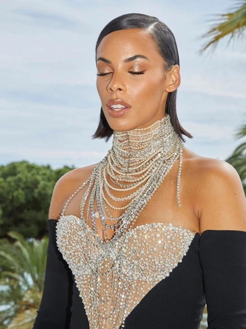 Close-up of a woman in an off-the-shoulder gown with layered rhinestone embellishments, slicked-back hair, and soft focus outdoor lighting.