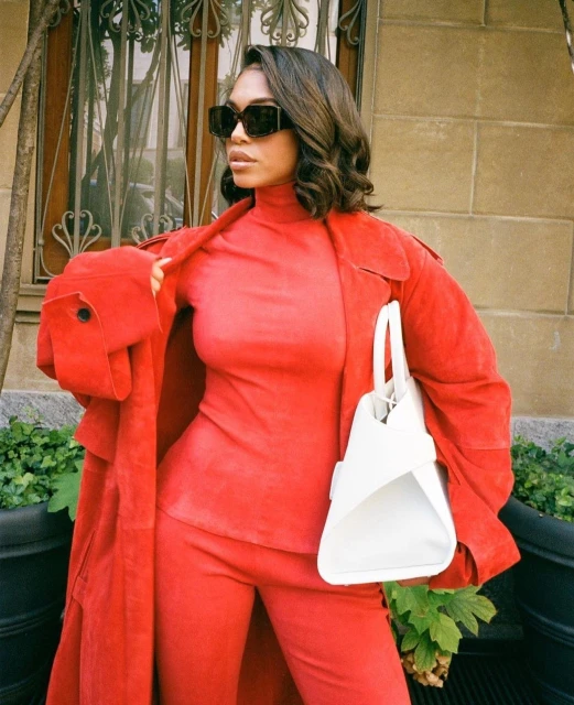 Woman in a red coat, turtleneck, and pants carrying a white purse on an outdoor city street with a neutral building background.
