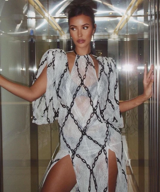 Woman poses in a white lace dress with black chain patterns in an elevator. She is wearing chandelier earrings and has a serious expression.