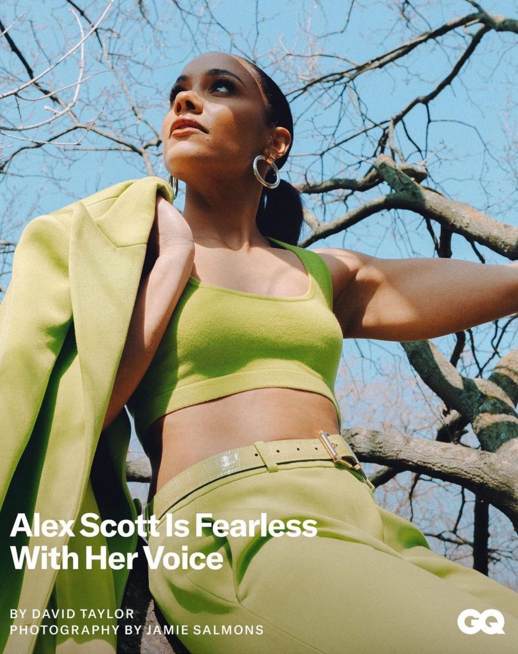 Alex Scott poses outdoors in a green outfit with a jacket, trees, and blue sky in the background. The image is an eye-level, medium shot.