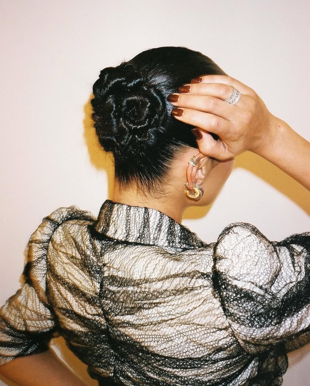 Woman with an elegant rose-shaped updo hairstyle, wearing a sheer black blouse and gold hoop earrings in a studio setting.