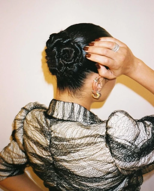 Woman with an elegant rose-shaped updo hairstyle, wearing a sheer black blouse and gold hoop earrings in a studio setting.