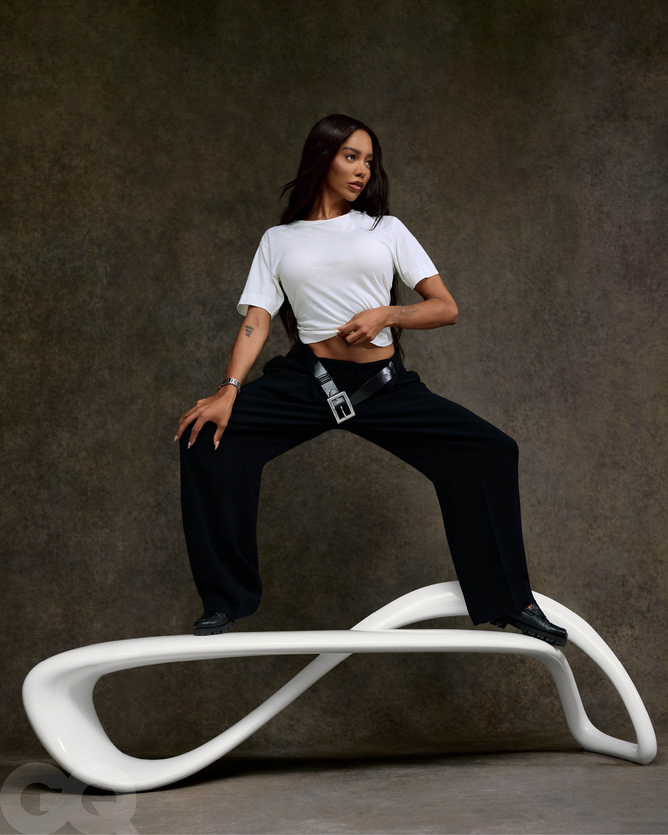 A young adult woman poses in a white t-shirt and black pants, standing on a white bench with a gray textured background for a studio fashion shot.