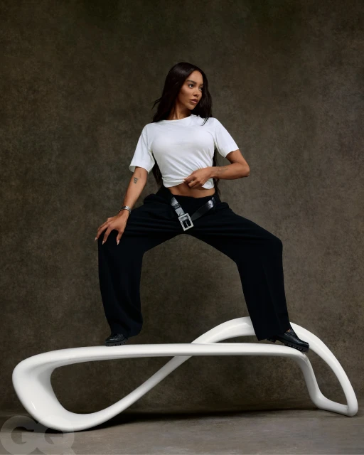 A young adult woman poses in a white t-shirt and black pants, standing on a white bench with a gray textured background for a studio fashion shot.