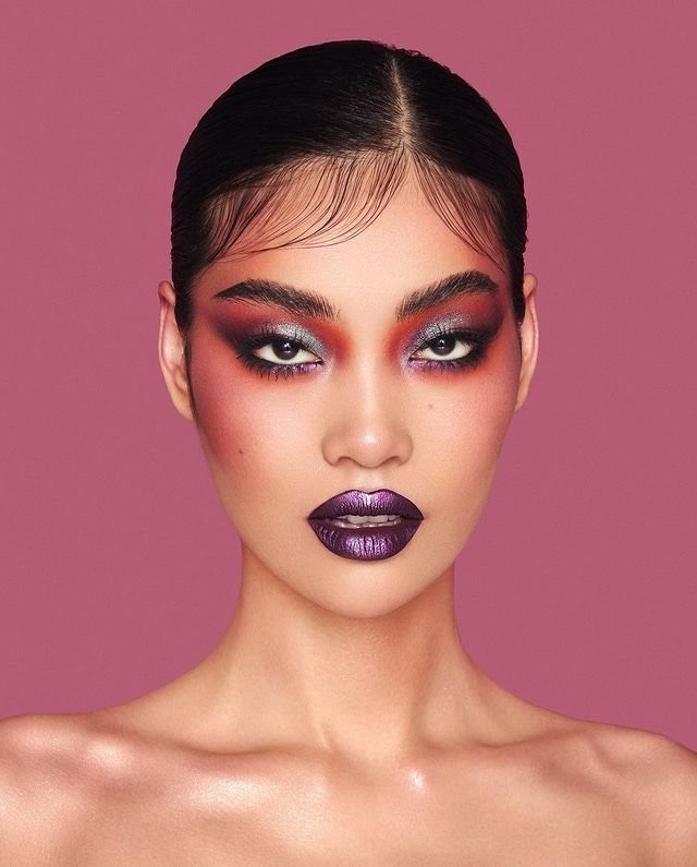 Headshot portrait of a young adult Asian woman with dramatic purple and red eye makeup and glossy purple lips against a pink background.