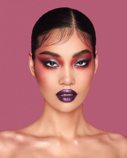 Headshot portrait of a young adult Asian woman with dramatic purple and red eye makeup and glossy purple lips against a pink background.