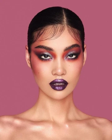 Headshot portrait of a young adult Asian woman with dramatic purple and red eye makeup and glossy purple lips against a pink background.