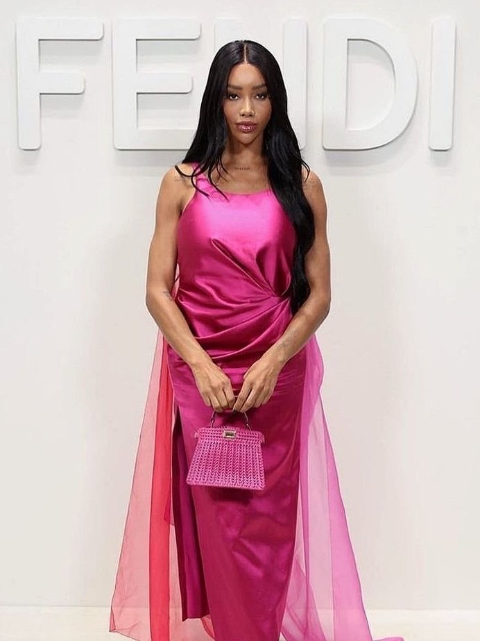Woman in fuchsia gown posing in front of a Fendi logo. She holds a matching woven handbag and has long, dark hair.