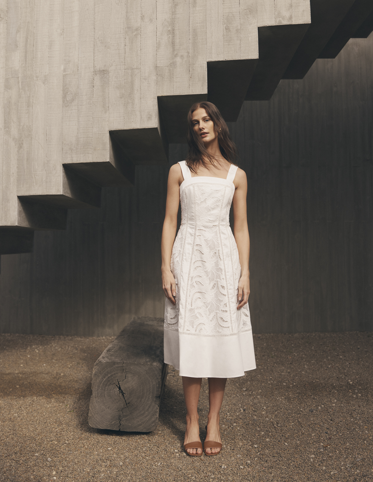 A woman wearing a white dress poses in front of a set of stairs and a textured wall, creating a minimalist and stylish fashion image.