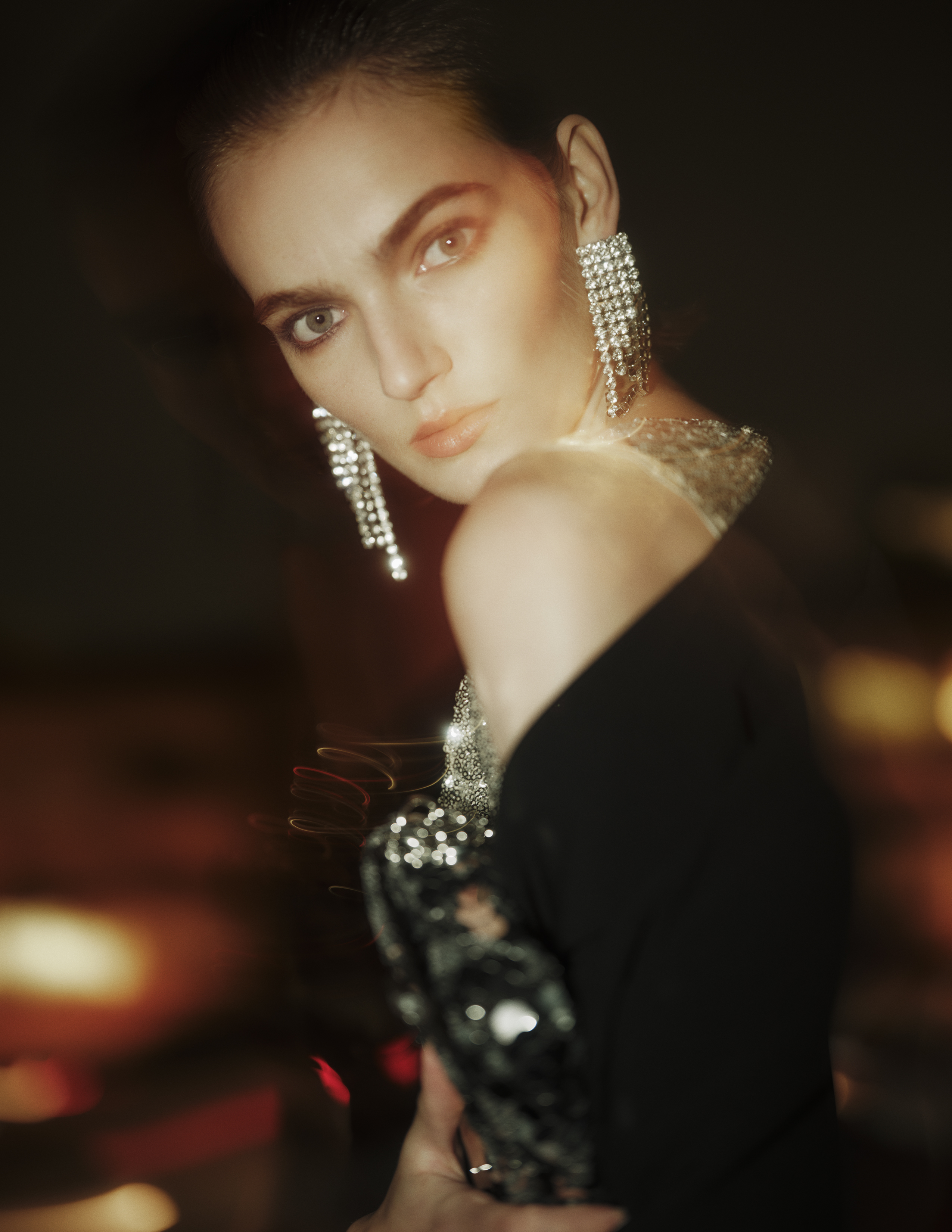Elegant woman in evening wear poses with sequined purse and cascading earrings against a dark background with bokeh lights.