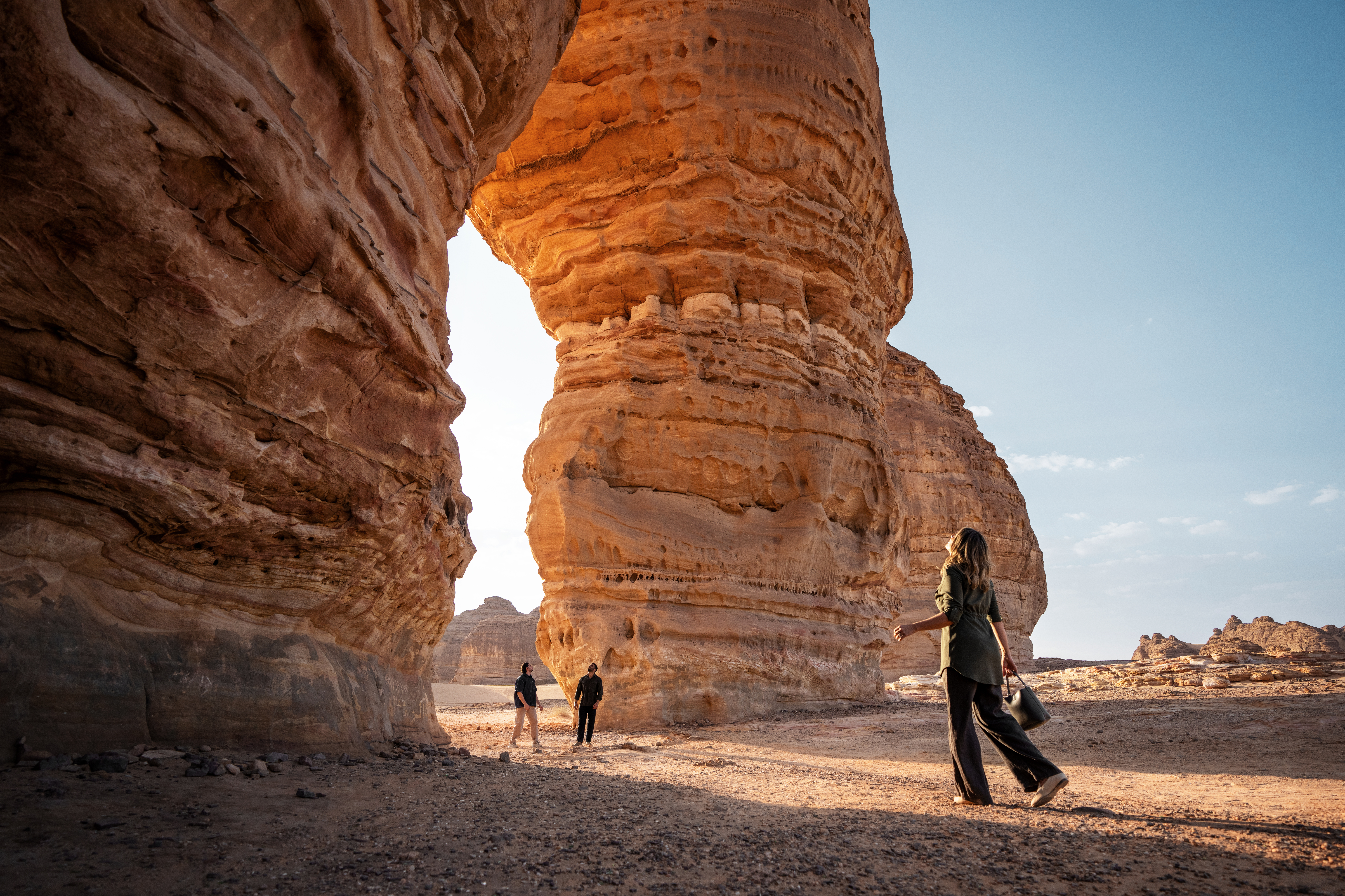 A woman walks near a large sandstone rock formation in a desert landscape under a clear sky, with two people standing in the background.