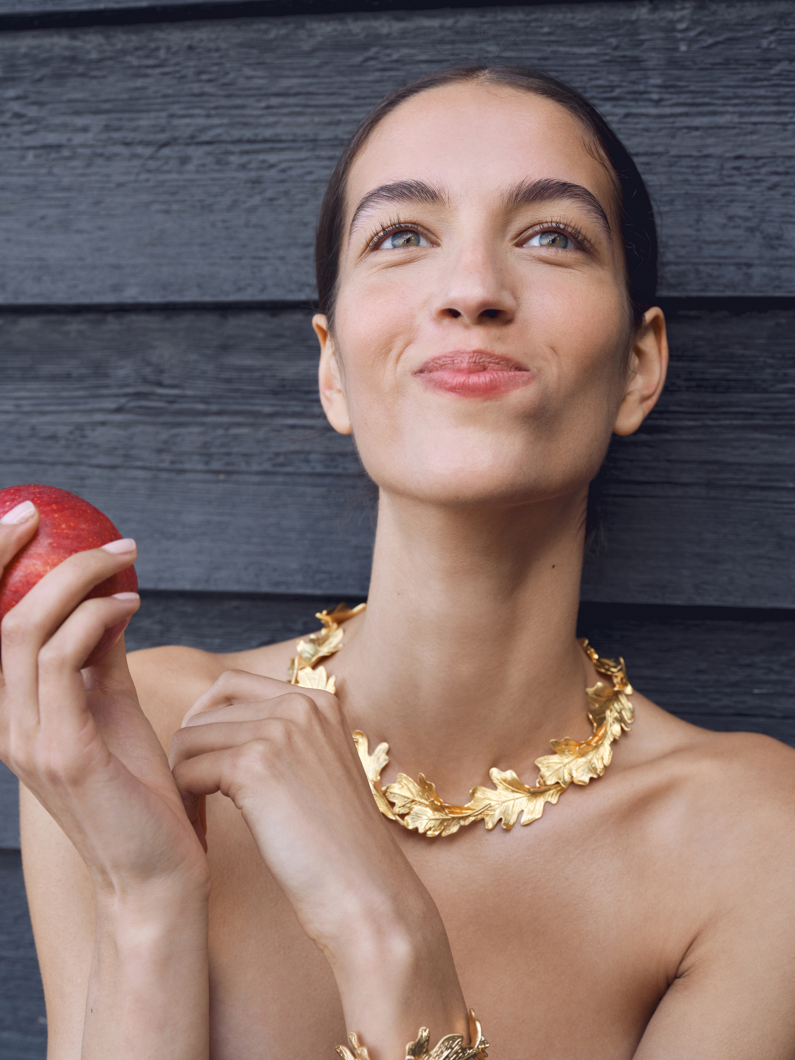 Close-up portrait of adult woman wearing leaf-inspired gold jewelry holding red apple in front of a dark gray wooden background.