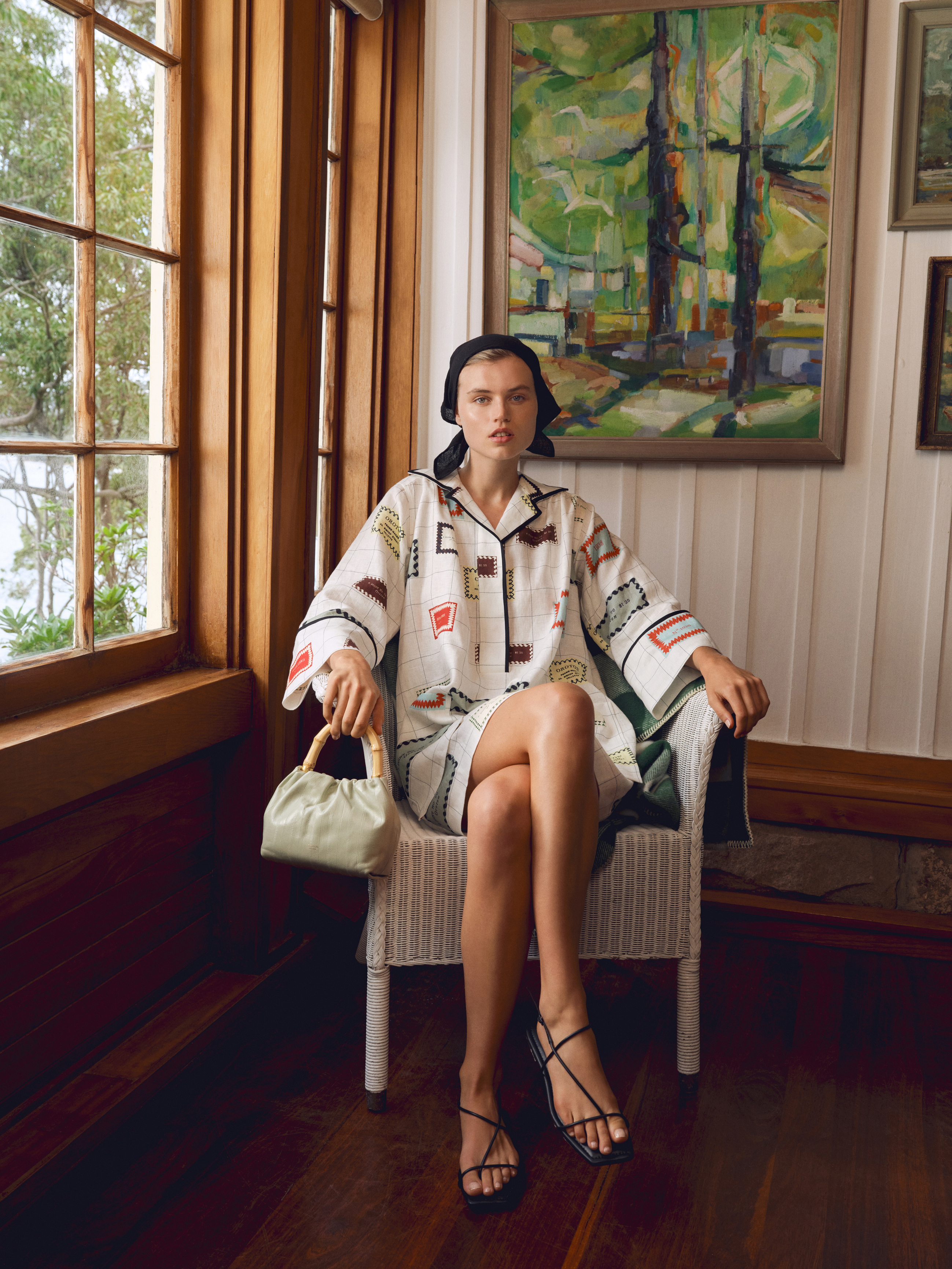 Indoor fashion shot of a fair-skinned woman in patterned pajamas sitting in a wicker chair with a small handbag and strappy sandals.