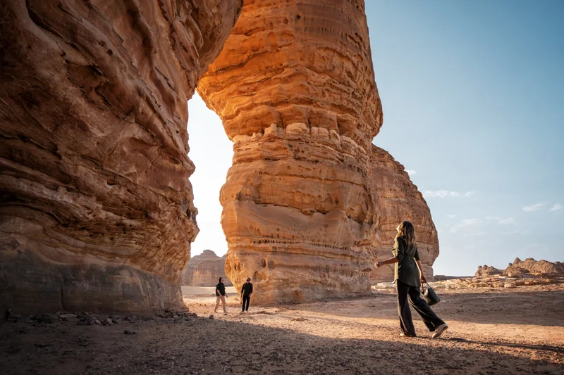 Woman in green shirt and dark pants standing in a desert landscape looking up at a massive natural sandstone arch with distant figures