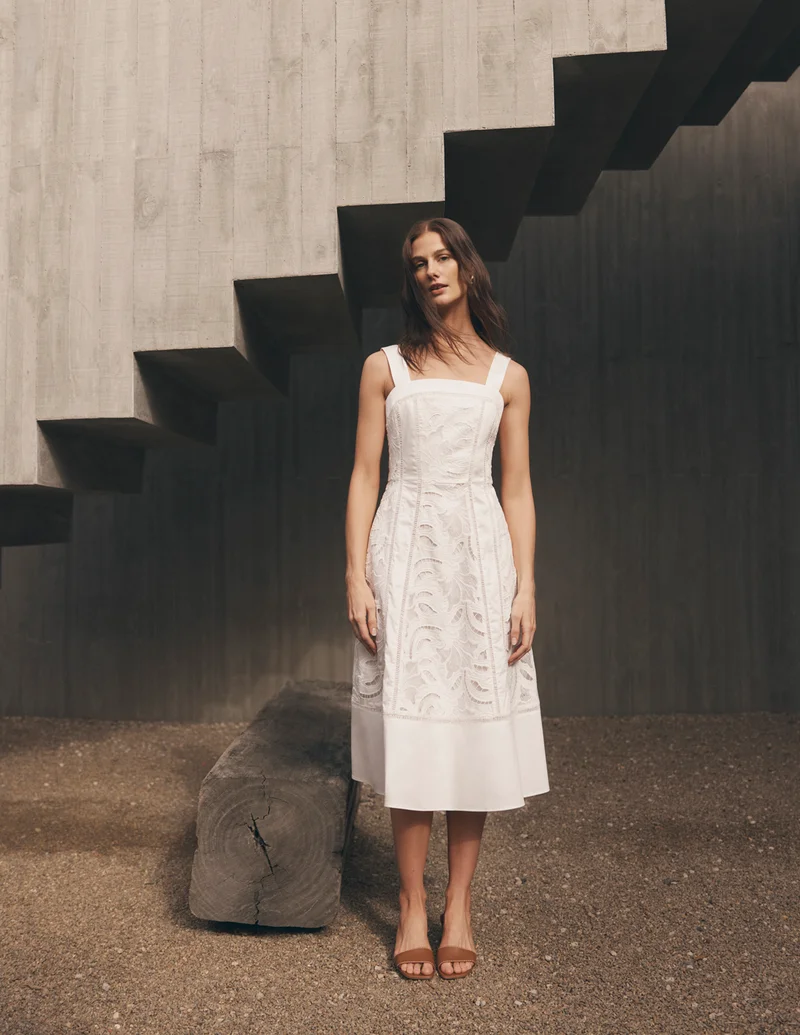 Woman in white midi dress standing before textured concrete staircase and gravel ground.