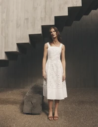 Woman in white midi dress standing before textured concrete staircase and gravel ground.