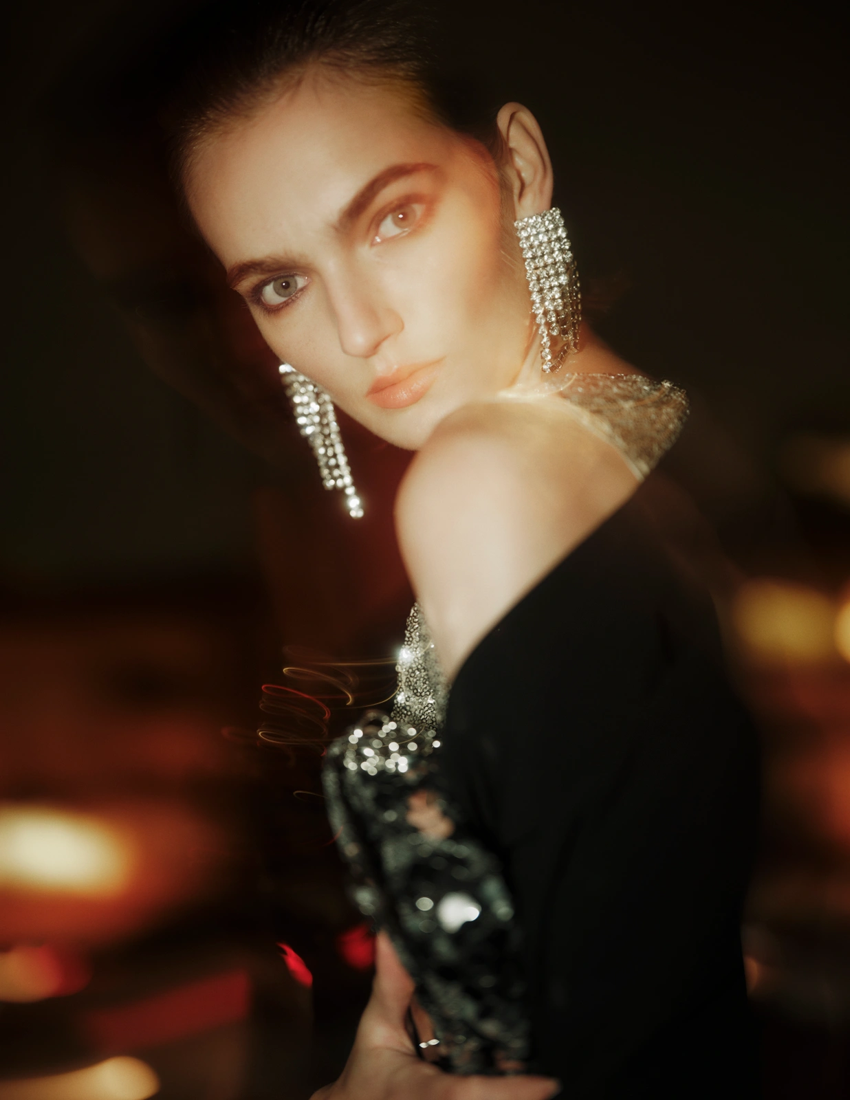 Woman looking over her bare shoulder wearing sparkling crystal fringe earrings and a black evening garment with warm blurred lighting.