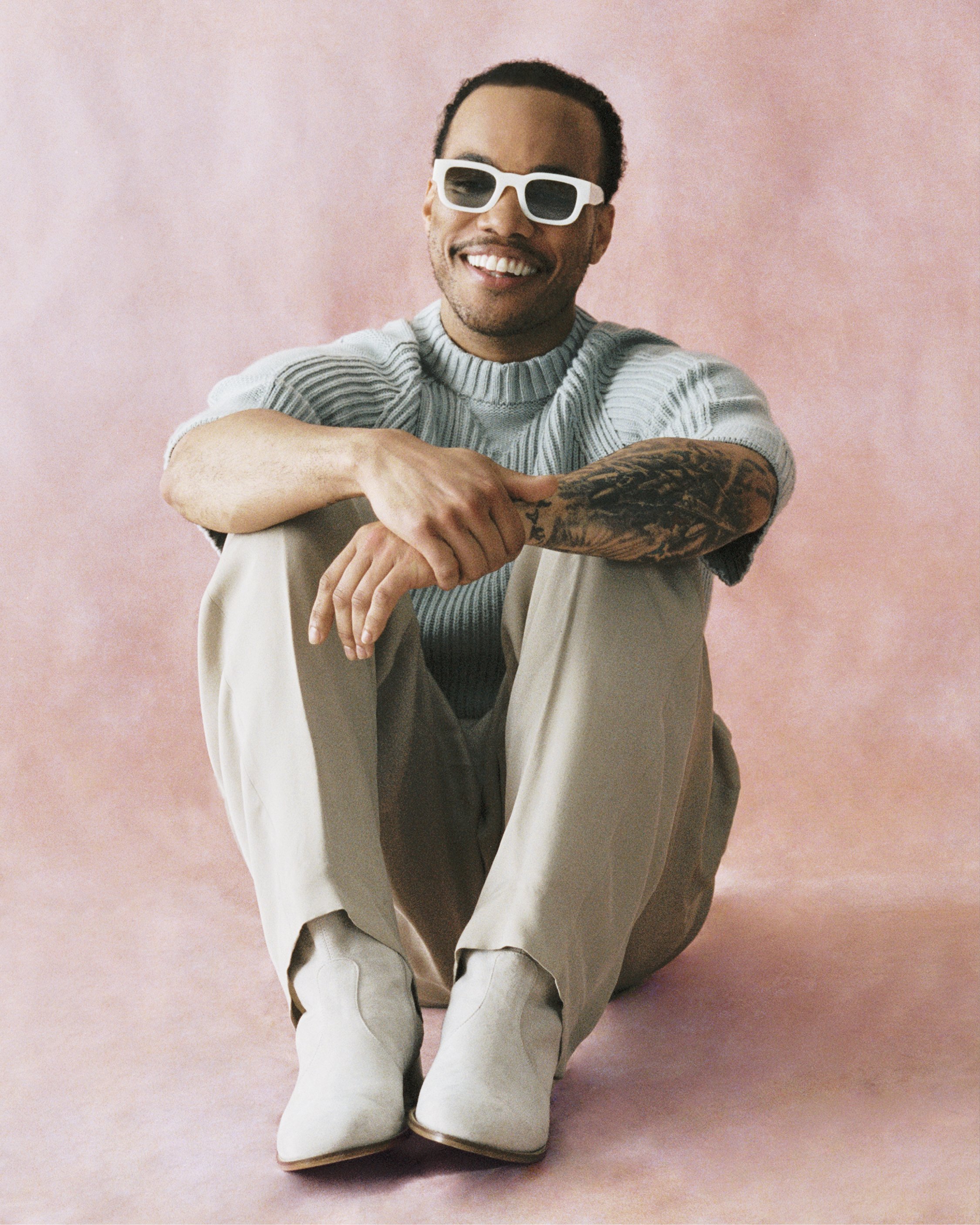 Smiling adult male in a light blue sweater and sunglasses poses in a studio with a pink backdrop and wears white boots.