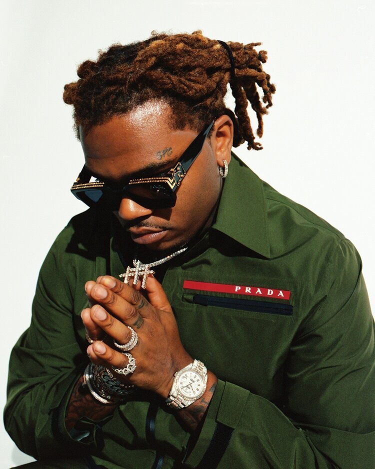 Adult male singer Gunna poses in a medium indoor shot wearing a green Prada jacket, diamond jewelry, and black sunglasses against a white background.