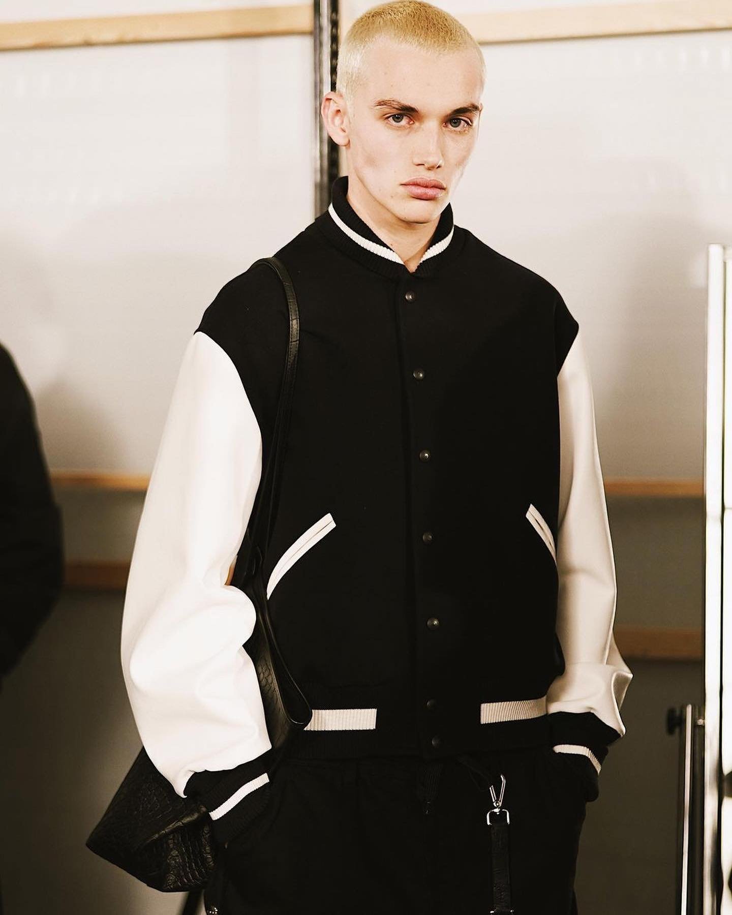 Standing adult male with short blonde hair poses in a black and white varsity jacket with a bag slung over his shoulder indoors.