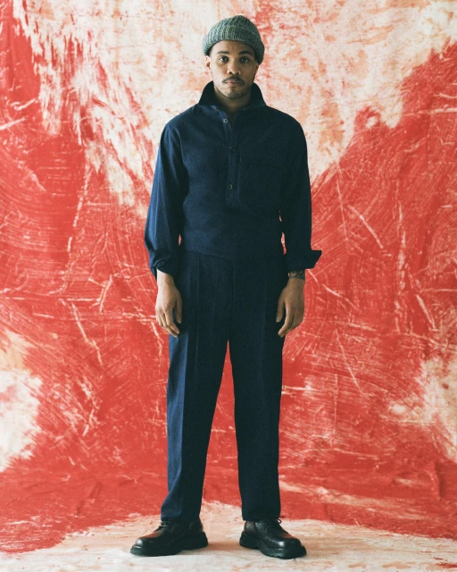 Man in dark blue shirt and trousers poses against a red-and-white background, wearing a knit hat and black leather shoes. Full shot.