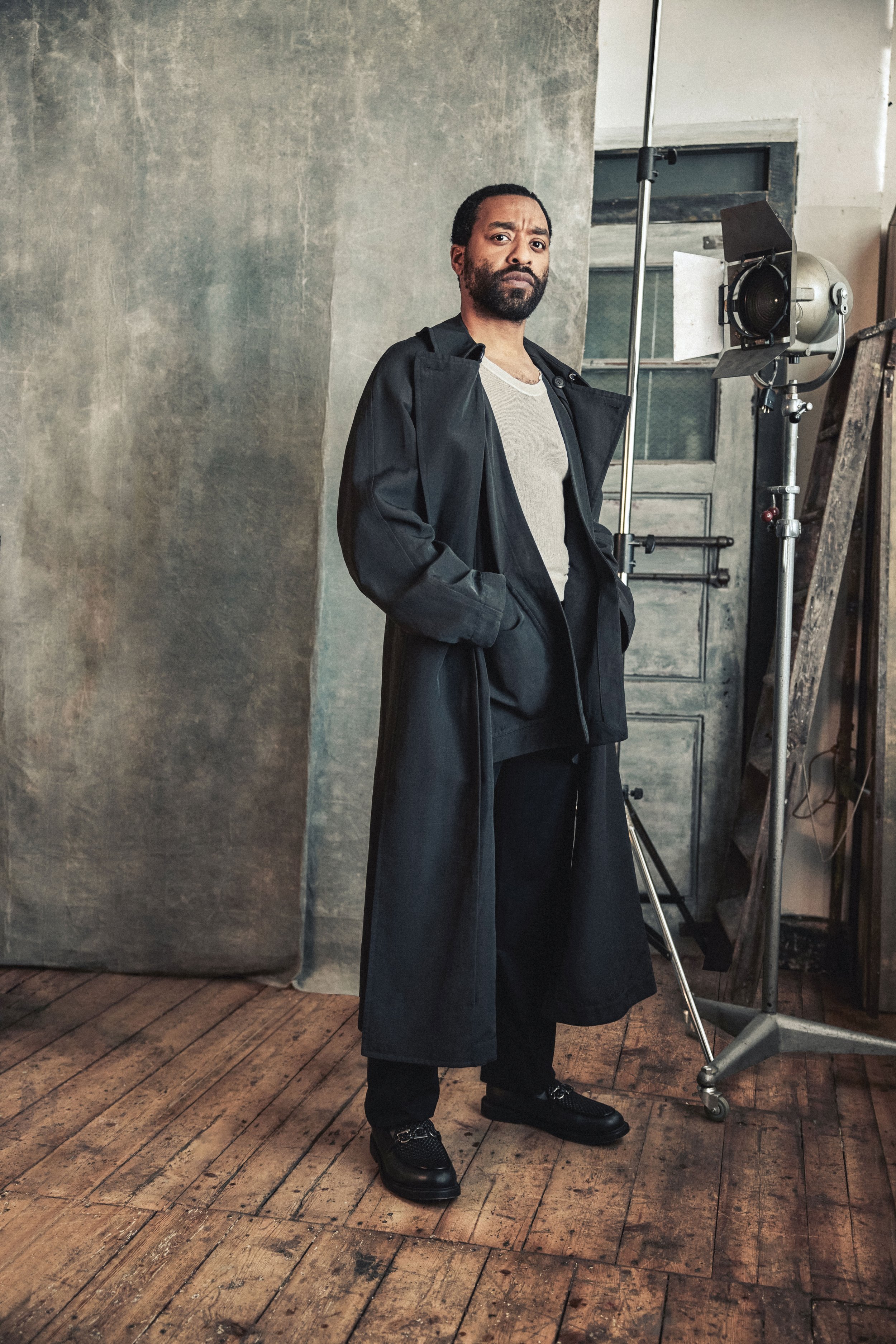 Portrait of an adult male in a long coat, standing in a photo studio against a textured backdrop with studio lighting.