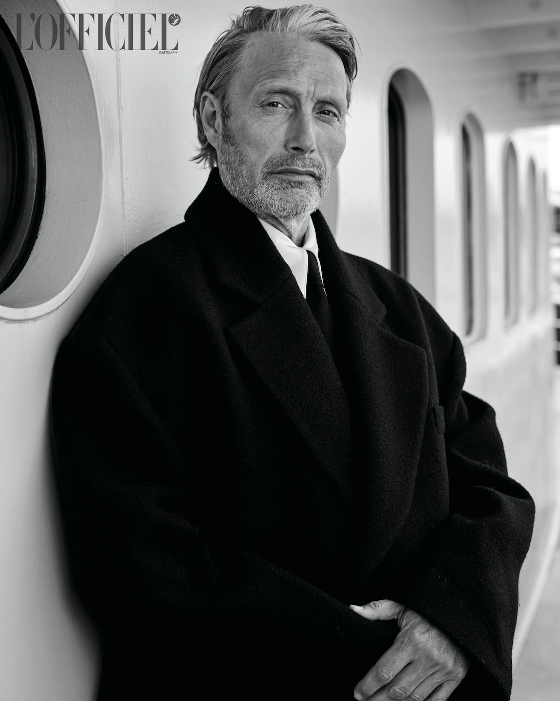 Monochrome portrait of Mads Mikkelsen in a dark coat leaning against a bright wall, shot at eye level in a well-lit indoor environment.