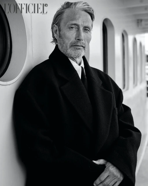 Monochrome portrait of Mads Mikkelsen in a dark coat leaning against a bright wall, shot at eye level in a well-lit indoor environment.